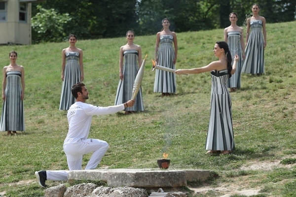 扮演最高女祭司的演員瑪麗·米娜（前右）點(diǎn)燃第一棒火炬手——希臘運(yùn)動(dòng)員、東京奧運(yùn)會(huì)男子賽艇項(xiàng)目金牌得主斯特凡諾斯·恩圖斯科斯手中的火炬。新華社記者 趙丁喆 攝